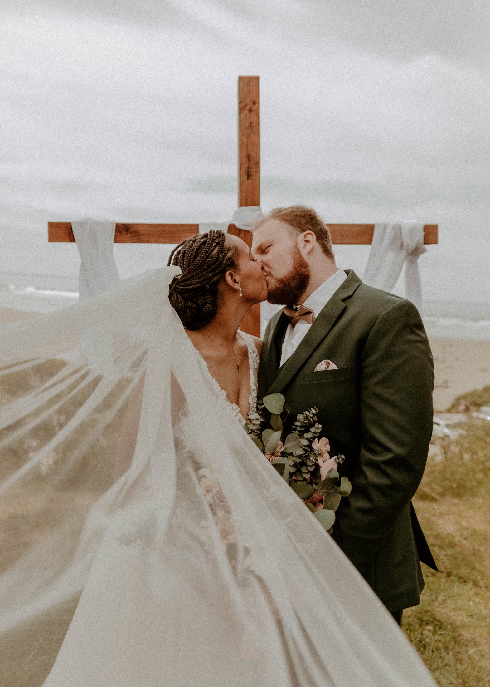 Danika and Grayson kissing at the driftwood cross altar with her veil blowing in the ocean wind